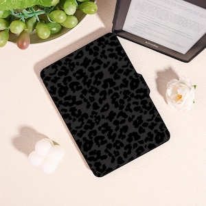 May include: Black Kindle case with a leopard print design. The case is rectangular and features a magnetic closure. The Kindle is visible in the background. The case is designed to protect the e-reader.