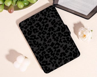May include: Black Kindle case with a leopard print design. The case is rectangular and features a magnetic closure. The Kindle is visible in the background. The case is designed to protect the e-reader.