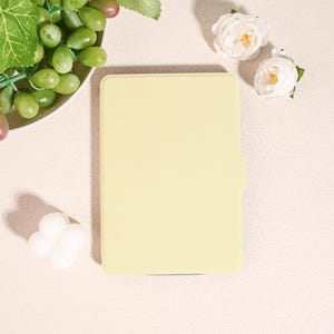 May include: A pale yellow tablet case with a smooth surface. The case is rectangular with rounded corners and a small tab on the right side. The image also includes white flowers, grapes, and a white decorative object.
