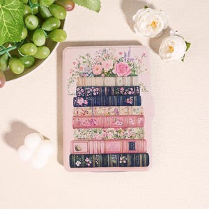 May include: A pink tablet case with a design of stacked books adorned with floral patterns. The books display diverse colours and patterns, including pink, blue, and floral prints. A bouquet of flowers, including pink roses, sits atop the stack.