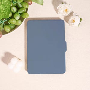 Könnte beinhalten: Eine schieferblaue Tablethülle mit Seitenlasche, auf einer cremefarbenen Oberfläche. Die Hülle ist rechteckig mit abgerundeten Ecken. Trauben und weiße Rosen sind um die Hülle angeordnet.