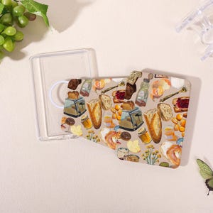 May include: A clear phone case with a breakfast-themed pattern. The design includes illustrations of toast, jam, honey, oranges, and a toaster on a beige background. The case is open, revealing the interior.