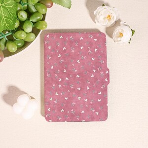 Può includere: Una custodia rettangolare rosa con un motivo floreale di piccoli cuori bianchi e foglie verdi. La custodia è chiusa e ha una chiusura magnetica. Lo sfondo include rose bianche e uva verde.