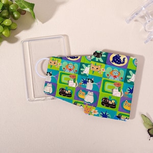 May include: A rectangular, foldable card case featuring a vibrant grid pattern of various illustrated cats in shades of green, blue, and purple. The design includes diverse feline poses, floral accents, and geometric shapes, with a clear plastic frame.