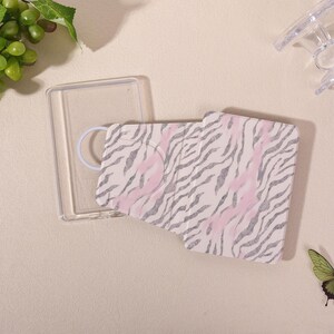May include: A rectangular clear plastic phone case with a magnetic ring attachment and a separate foldable cover featuring a pink and silver zebra stripe pattern. The cover has a cutout for a camera lens and a textured surface design.
