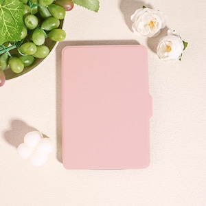 May include: A pale pink e-reader case with a smooth finish. The case is rectangular with rounded corners and a side flap. The background includes white roses and a plate of green grapes.