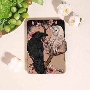 Op de afbeelding: Een decoratieve tablethoes met een vintage illustratie van een zwarte raaf en een witte uil op een bloeiende tak. Het ontwerp bevat roze bloesems en een neutrale achtergrond. De hoes is ontworpen om een tablet te beschermen.