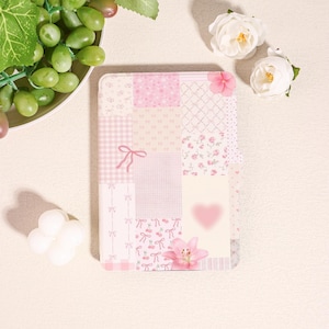 May include: A rectangular, patchwork-style notebook cover in shades of pink. The design includes various patterns like gingham, bows, and floral prints. A pink flower and a heart accent the cover. The notebook is surrounded by white flowers and grapes.