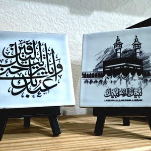 May include: Two square ceramic tiles on small black easels. One tile features black Arabic calligraphy, the other a black and white illustration of a building with text. A black decorative box is in the background.