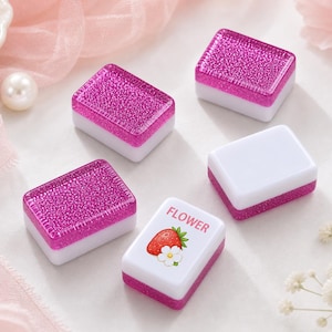 May include: Five rectangular mahjong tiles with a two-tone design. Four tiles feature a solid white base with a vibrant, glittery magenta top. One tile has a white base with a magenta bottom and a graphic of a red strawberry and white flower with the text FLOWER.