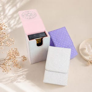 May include: Three glittery card cases in white, purple, and silver. The open silver case reveals a deck of cards. The purple case has a dotted pattern. The open case has a pink interior and a silver emblem on the lid. The cases are for tarot cards.