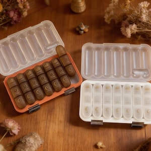 May include: Two open pill organizers with clear, rectangular compartments. One is orange with brown pill containers, the other is white. Each container has a hinged lid and is designed for daily medication management.