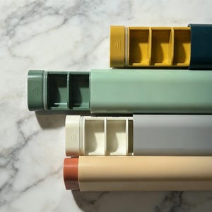 Puede incluir: Vista cenital de cinco pastilleros rectangulares de colores. Cada pastillero tiene una tapa corredera y un interior compartimentado. Los colores incluyen amarillo, verde azulado, verde claro, gris y beige. Los pastilleros están dispuestos en diagonal sobre una superficie de mármol.