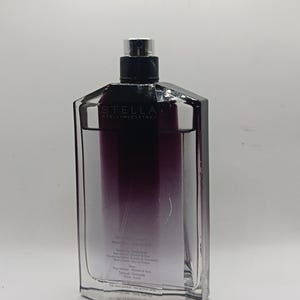 May include: A rectangular perfume bottle with a black cap and a gradient of purple to clear liquid. The bottle has the brand name "STELLA" printed on the front.