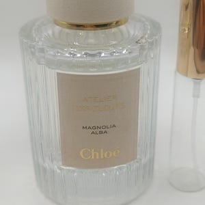 Pode incluir: Um frasco de perfume de vidro transparente com tampa bege e detalhes dourados. O rótulo diz "ATELIER DES FLEURS MAGNOLIA ALBA Chloe". Ao lado, um frasco de vidro menor e vazio com tampa dourada.