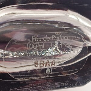May include: Close-up of a clear perfume bottle with the text "Eau de Parfum" and "100 ml - 3.3 fl. oz." The brand name "Stella McCartney Parfums" is also visible. The bottle has a reflective, glossy surface.