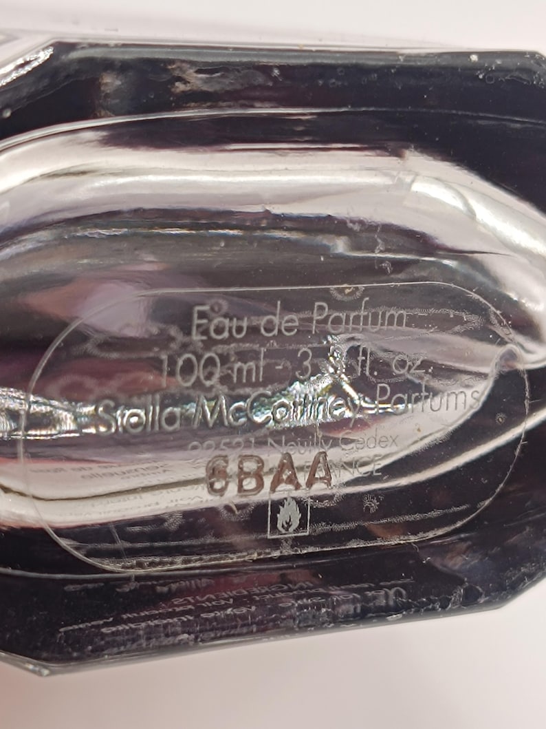 May include: Close-up of a clear perfume bottle with the text "Eau de Parfum" and "100 ml - 3.3 fl. oz." The brand name "Stella McCartney Parfums" is also visible. The bottle has a reflective, glossy surface.