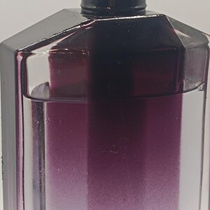 May include: A close-up of a perfume bottle with a dark purple gradient. The bottle has a geometric design with a black cap and a silver atomizer. The liquid inside transitions from clear to a deep purple.