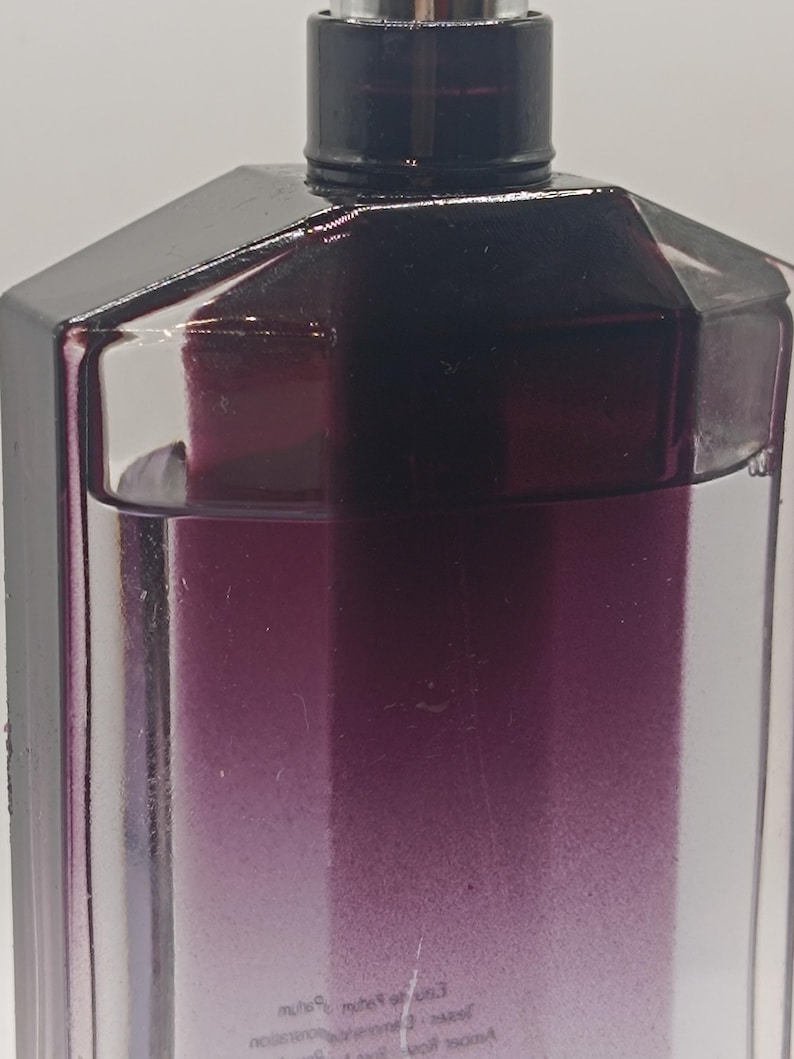 May include: A close-up of a perfume bottle with a dark purple gradient. The bottle has a geometric design with a black cap and a silver atomizer. The liquid inside transitions from clear to a deep purple.