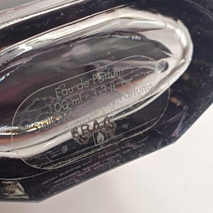 May include: A clear glass perfume bottle with a dark, angular design. The label reads "Eau de Parfum 100 ml - 3.3 fl oz" and includes the brand name "Stella McCartney Parfums". The bottle is partially visible, with a focus on the label.