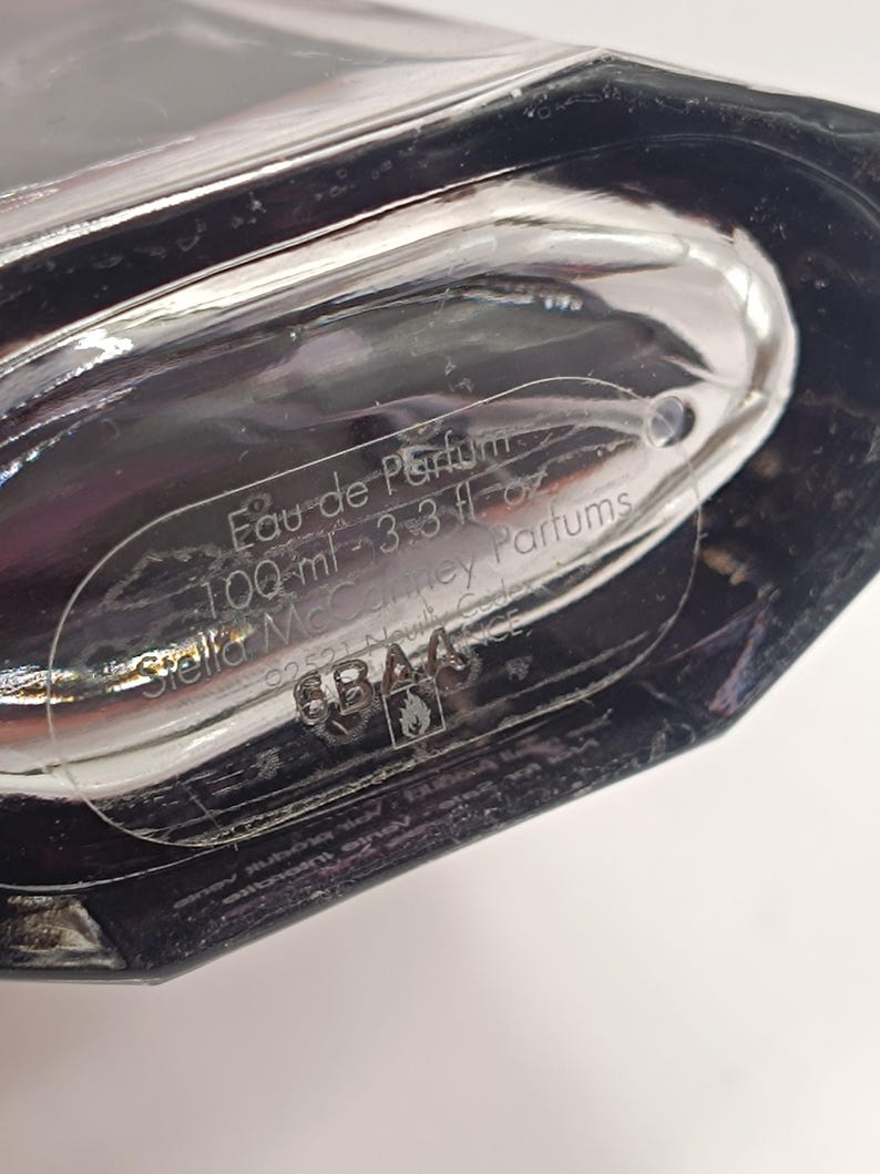 May include: A clear glass perfume bottle with a dark, angular design. The label reads "Eau de Parfum 100 ml - 3.3 fl oz" and includes the brand name "Stella McCartney Parfums". The bottle is partially visible, with a focus on the label.