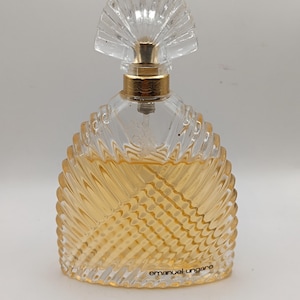Puede incluir: Un frasco de perfume de vidrio transparente con un tapón en forma de abanico y detalles dorados. La botella tiene un diseño texturizado y acanalado y contiene un líquido ámbar claro. El texto "emanuel ungaro" está impreso en la parte inferior del frasco.