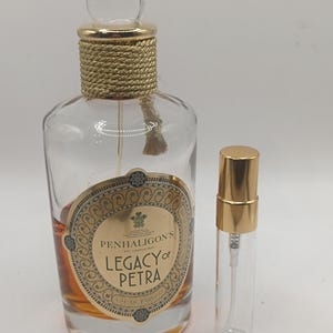 Pode incluir: Um frasco de perfume de vidro transparente com uma tampa dourada e um frasco de amostra menor. O frasco maior tem um rótulo dourado com o texto "PENHALIGON'S LEGACY of PETRA". O frasco está cheio de um líquido castanho claro.