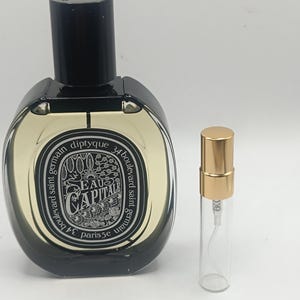 Pode incluir: Um frasco de perfume preto e transparente com uma tampa preta e um frasco de amostra menor com uma tampa dourada. O frasco de perfume tem um rótulo que diz "EAU CAPITALE" e "diptyque 34 boulevard saint germain paris 5e".