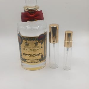 Pode incluir: Um frasco de perfume de vidro transparente com um rótulo dourado e uma fita vermelha, acompanhado por dois frascos de spray menores. O rótulo diz "Penhaligon's Constantinople Eau de Parfum 100ml 3.4 FL OZ."