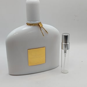 Op de afbeelding: Een witte parfumfles met een geribbelde dop en een goudkleurig accent. Een kleinere, transparante monsterfles met een zilveren dop staat ernaast. De parfumfles heeft een gouden label met tekst.