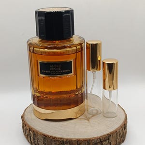 Pode incluir: Um frasco de perfume e dois atomizadores menores estão em uma fatia de madeira. O frasco grande é âmbar com uma tampa preta e base dourada. O rótulo diz "AMBER DESIRE CAROLINA HERRERA NEW YORK". Os frascos menores têm tampas douradas.