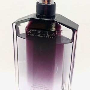 May include: A clear, rectangular perfume bottle with a black cap and a silver sprayer. The bottle contains a purple-to-clear gradient liquid. The brand name "STELLA McCARTNEY" is printed on the front.