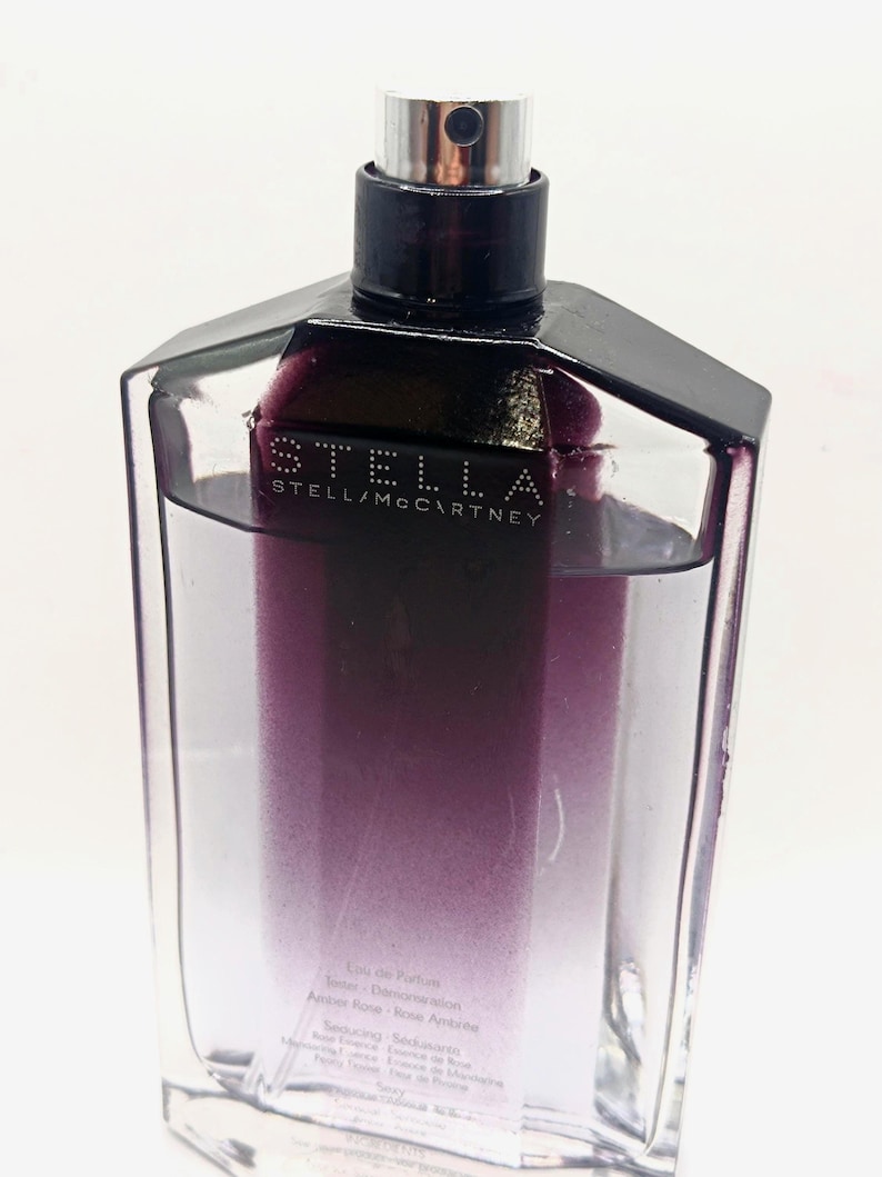 May include: A clear, rectangular perfume bottle with a black cap and a silver sprayer. The bottle contains a purple-to-clear gradient liquid. The brand name "STELLA McCARTNEY" is printed on the front.