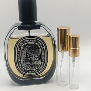 Pode incluir: Um frasco de perfume oval transparente com tampa preta e rótulo dourado, ao lado de dois pequenos frascos de spray de vidro com tampas douradas. O frasco de perfume tem o texto "EAU DUELLE" e "diptyque" no rótulo.
