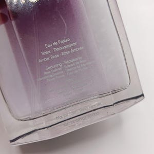 May include: A clear perfume bottle with a purple liquid inside. The bottle is labeled "Eau de Parfum" and "Tester - D&eacute;monstration". The text also includes "Amber Rose - Rose Ambr&eacute;e" and "Seducing - S&eacute;duisante". The bottom of the bottle reads "Not for Sale - Vente interdite".