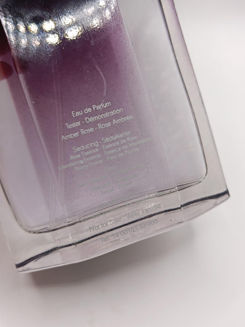 May include: A clear perfume bottle with a purple liquid inside. The bottle is labeled "Eau de Parfum" and "Tester - D&eacute;monstration". The text also includes "Amber Rose - Rose Ambr&eacute;e" and "Seducing - S&eacute;duisante". The bottom of the bottle reads "Not for Sale - Vente interdite".