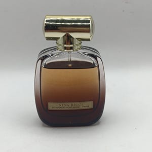 Könnte beinhalten: Ein Parfümflakon mit einem transparenten, quadratischen Glaskörper und einem Farbverlauf aus brauner Flüssigkeit im Inneren. Die Flasche hat einen goldfarbenen Verschluss und ein Etikett mit der Aufschrift "NINA RICCI 39 AVENUE MONTAIGNE - PARIS".