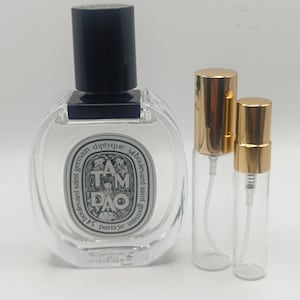 Pode incluir: Um frasco de perfume transparente, de forma oval, com uma tampa preta, ladeado por dois frascos de vidro menores com borrifadores e tampas douradas. O frasco de perfume tem um rótulo com o texto "TAM DAO" e "diptyque".