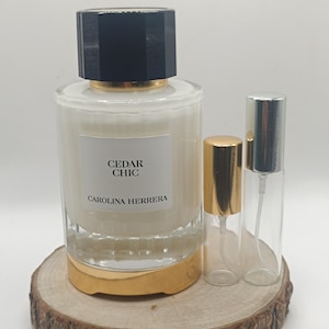 Może przedstawiać: Flakon perfum Carolina Herrera Cedar Chic z białą cieczą, czarną nakrętką i złotą podstawą, w towarzystwie dwóch małych, przezroczystych szklanych fiolek z rozpylaczem ze złotymi i srebrnymi nakrętkami, umieszczonych na okrągłej drewnianej podstawie.