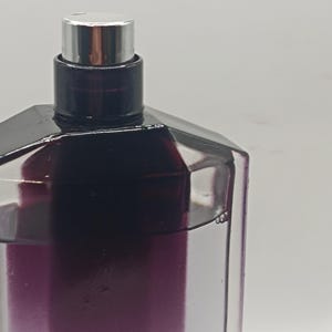 May include: A close-up of a perfume bottle with a geometric design. The bottle is clear with a gradient of purple hues. It has a black cap and a silver spray nozzle. The background is a plain white.