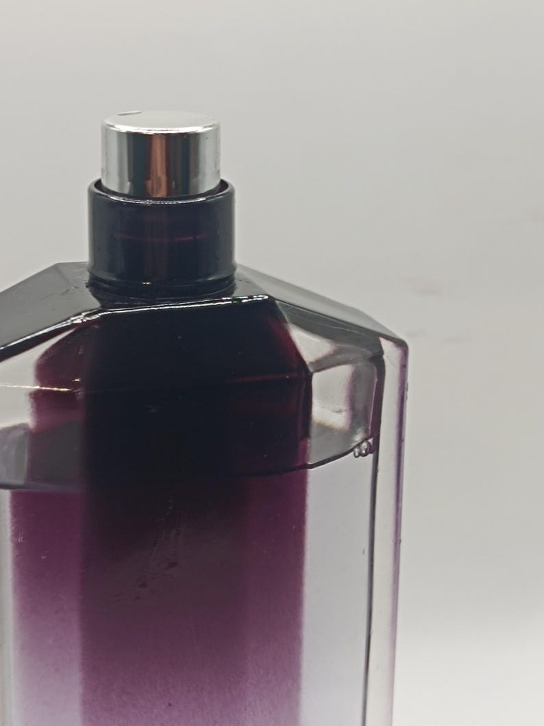 May include: A close-up of a perfume bottle with a geometric design. The bottle is clear with a gradient of purple hues. It has a black cap and a silver spray nozzle. The background is a plain white.