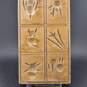 Könnte beinhalten: Eine rechteckige, hellbraune Holzform mit sechs verschiedenen geschnitzten Abschnitten. Jeder Abschnitt enthält ein einzigartiges vertieftes Design, darunter stilisierte Blumen-, Tier- und Pflanzenmotive, mit sichtbarer Holzmaserung und glatten Kanten.