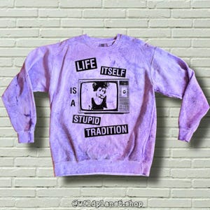 May include: A light purple sweatshirt with a tie-dye effect. The front features a black and white graphic of a person on a vintage television screen. The text "LIFE IS A STUPID TRADITION" is printed around the television.
