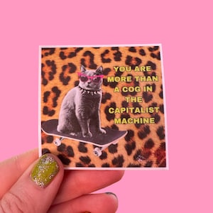 May include: A gray cat wearing pink sunglasses and a silver choker is skateboarding on a leopard print background. The text "You are more than a cog in the capitalist machine" is written in yellow on a white background.