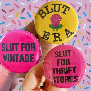May include: Three round buttons with bold text. One button is yellow with "SLUT ERA" and a diamond ring graphic. Two buttons are pink, one with "SLUT FOR VINTAGE" and the other with "SLUT FOR THRIFT STORES".