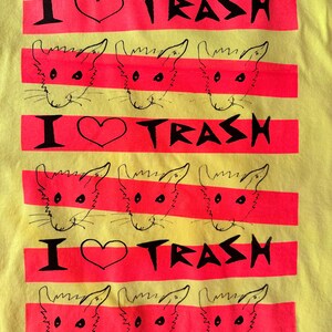 May include: Yellow t-shirt with a graphic design featuring the text "I ❤️ TRASH" in black, with a heart symbol, and a series of red horizontal stripes. Each stripe contains a cartoon cat face.
