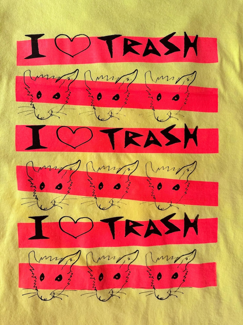 May include: Yellow t-shirt with a graphic design featuring the text "I ❤️ TRASH" in black, with a heart symbol, and a series of red horizontal stripes. Each stripe contains a cartoon cat face.
