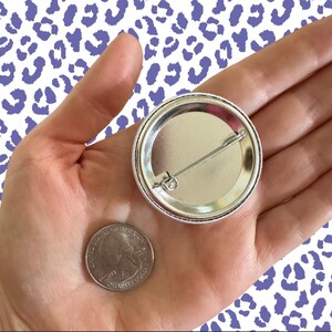 Peut inclure: Un bouton argent&eacute; avec une &eacute;pingle, tenu dans une main &agrave; c&ocirc;t&eacute; d'un quart de dollar am&eacute;ricain. Le bouton a un bord en m&eacute;tal brillant et un centre vierge de couleur claire. Le fond pr&eacute;sente un motif blanc avec des taches de l&eacute;opard violettes.