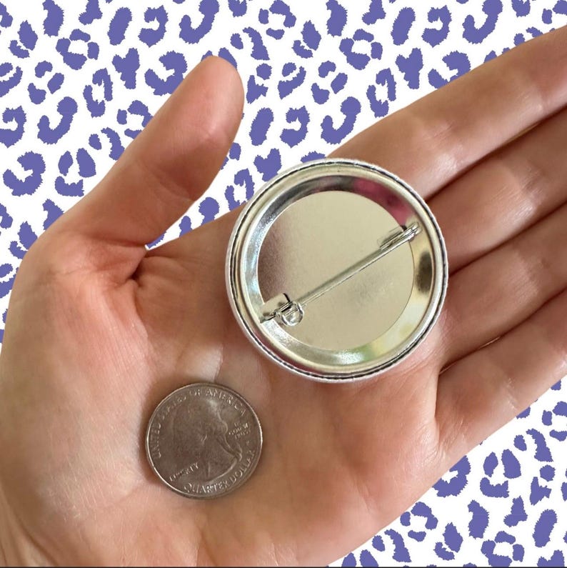 Peut inclure: Un bouton argent&eacute; avec une &eacute;pingle, tenu dans une main &agrave; c&ocirc;t&eacute; d'un quart de dollar am&eacute;ricain. Le bouton a un bord en m&eacute;tal brillant et un centre vierge de couleur claire. Le fond pr&eacute;sente un motif blanc avec des taches de l&eacute;opard violettes.