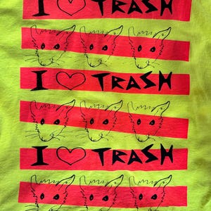 Pu&ograve; includere: T-shirt giallo neon con un design grafico. Il design presenta il testo "I ❤️ TRASH" in nero su una striscia orizzontale rossa, alternato a facce di gatti dei cartoni animati. La maglietta &egrave; audace e accattivante.